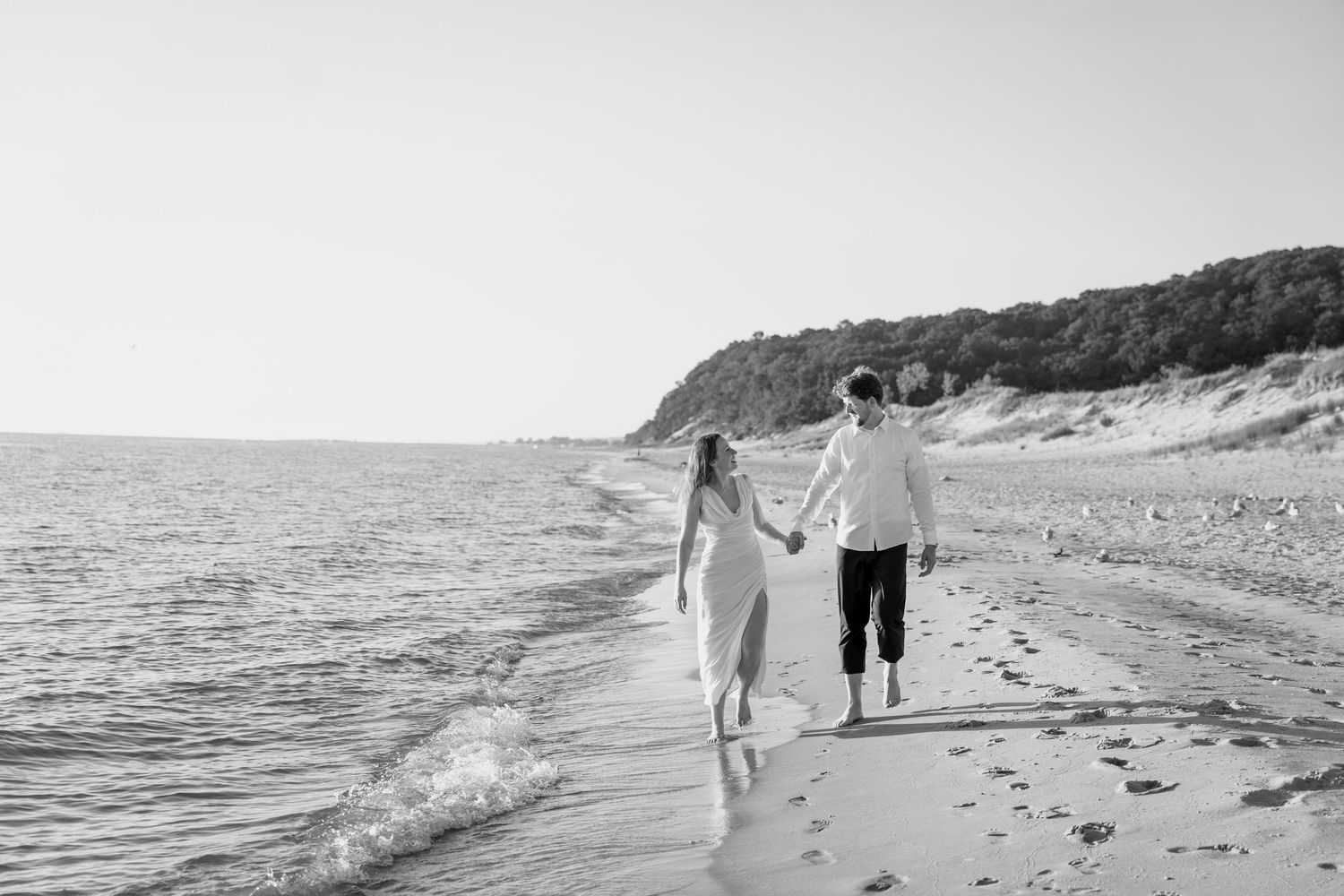 Black and white photograph of a couple strolling hand in hand by the ocean.