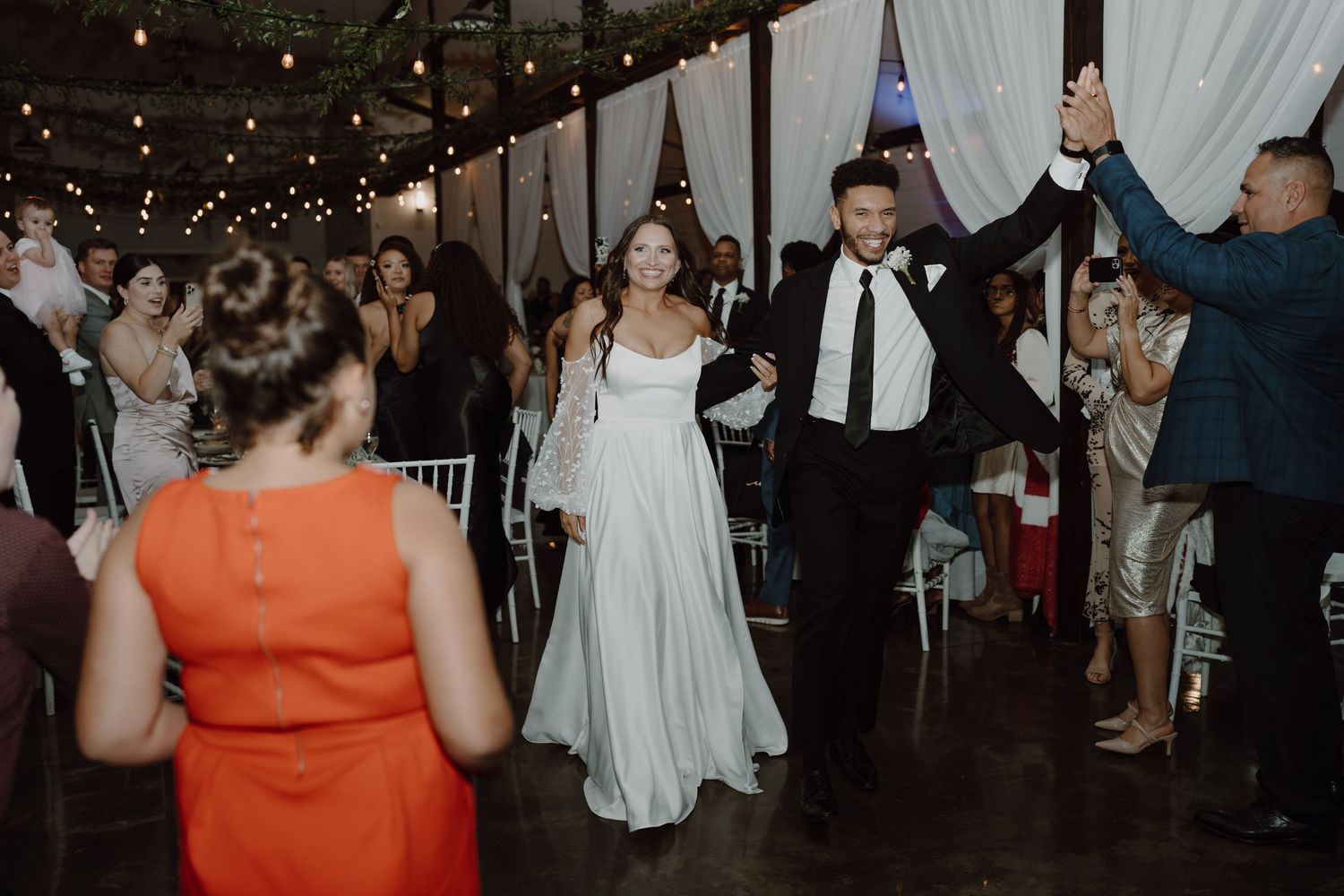 Wedding celebration in progress at an indoor reception venue with string lights and white decor.
