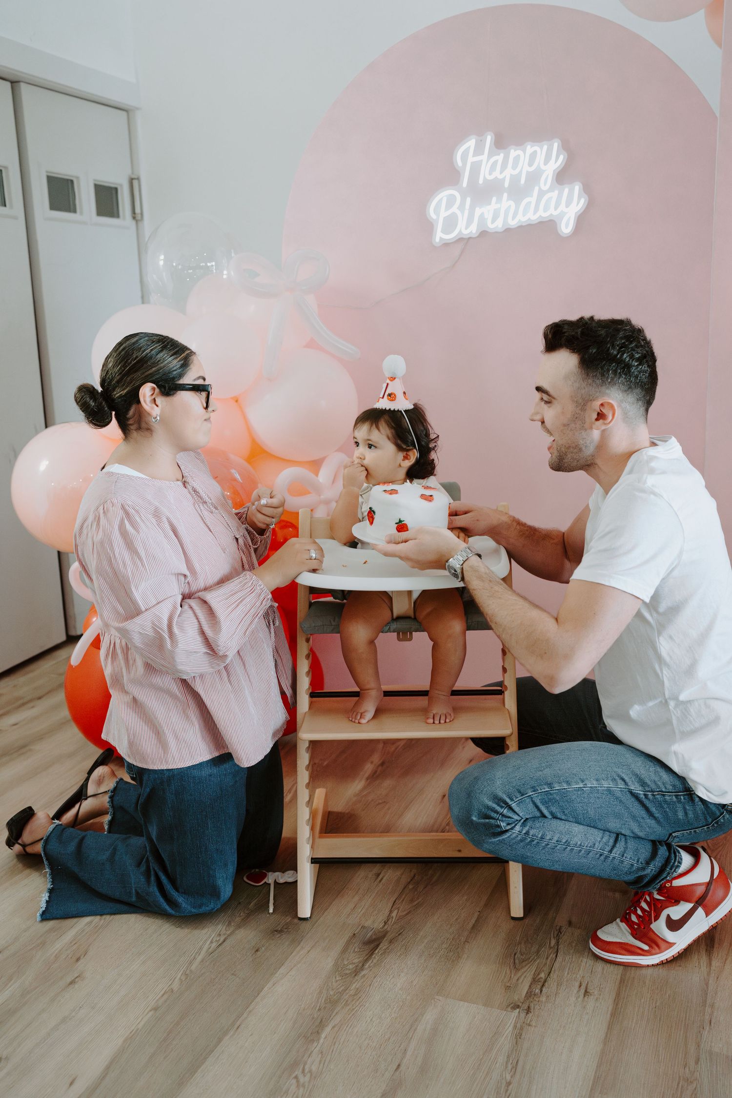 Sweet birthday moment captured with family around small table against pink balloon backdrop.