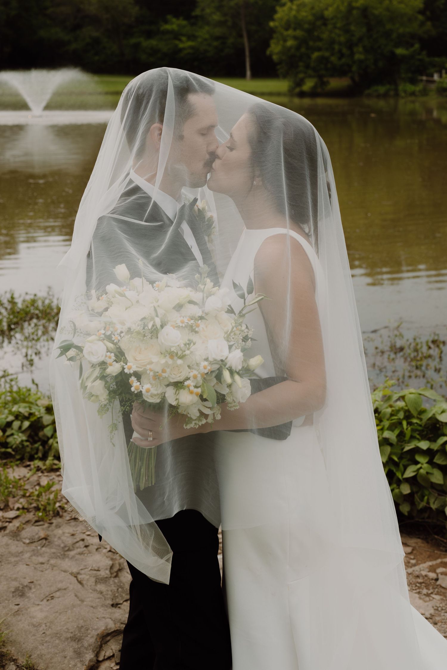 Romantic lakeside wedding scene with flowing veil and white floral bouquet.