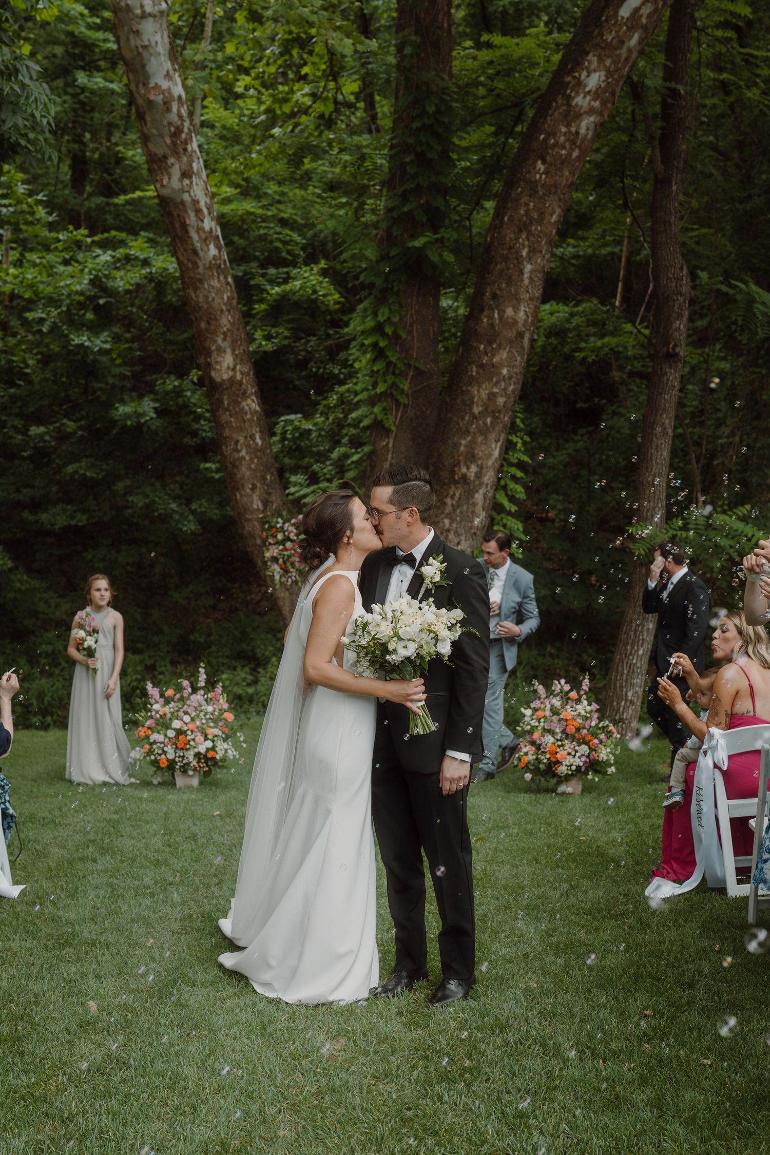 A wedding ceremony takes place in a wooded outdoor setting with white chairs and floral arrangements.