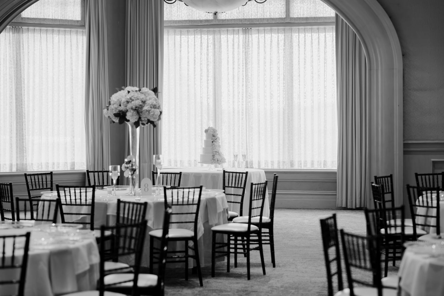 Black and white photograph of an elegant reception hall with rows of empty chairs and tall windows.