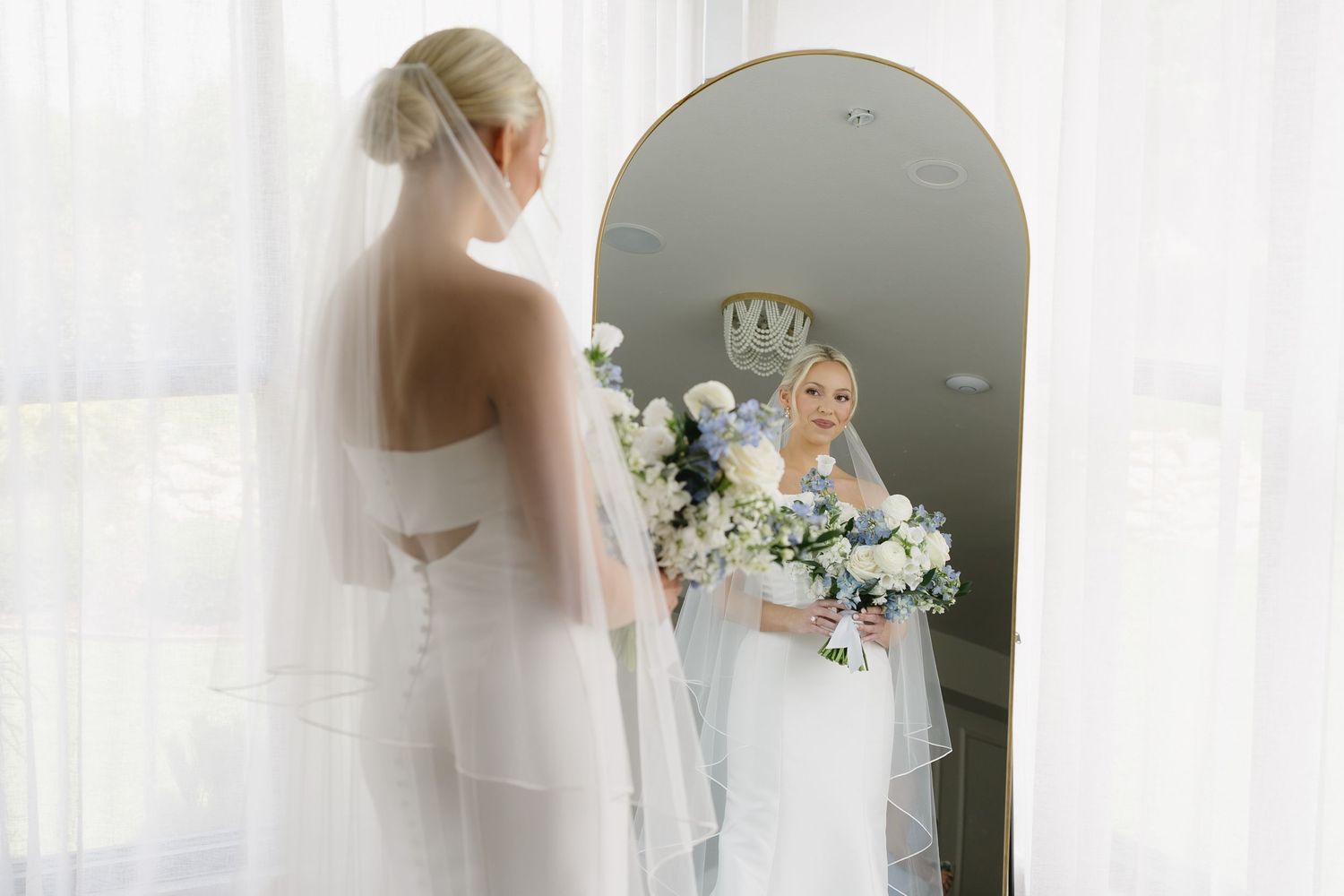 Bride in white gown and veil holds bouquet of white and blue flowers, admiring her reflection in an arched mirror.