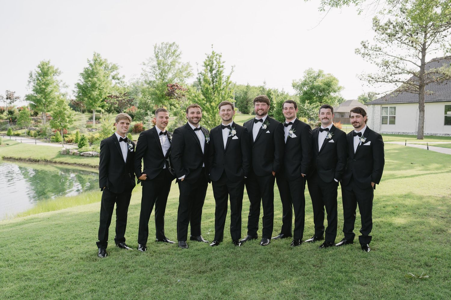 Groom and groomsmen in black tuxedos with bow ties posing outdoors on green lawn near pond at wedding.