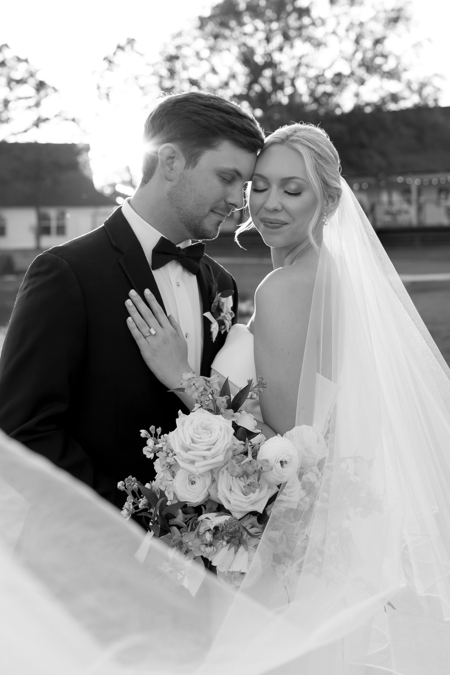 Black and white photo of bride and groom embracing, bride holding floral bouquet with veil flowing, outdoor wedding portrait.