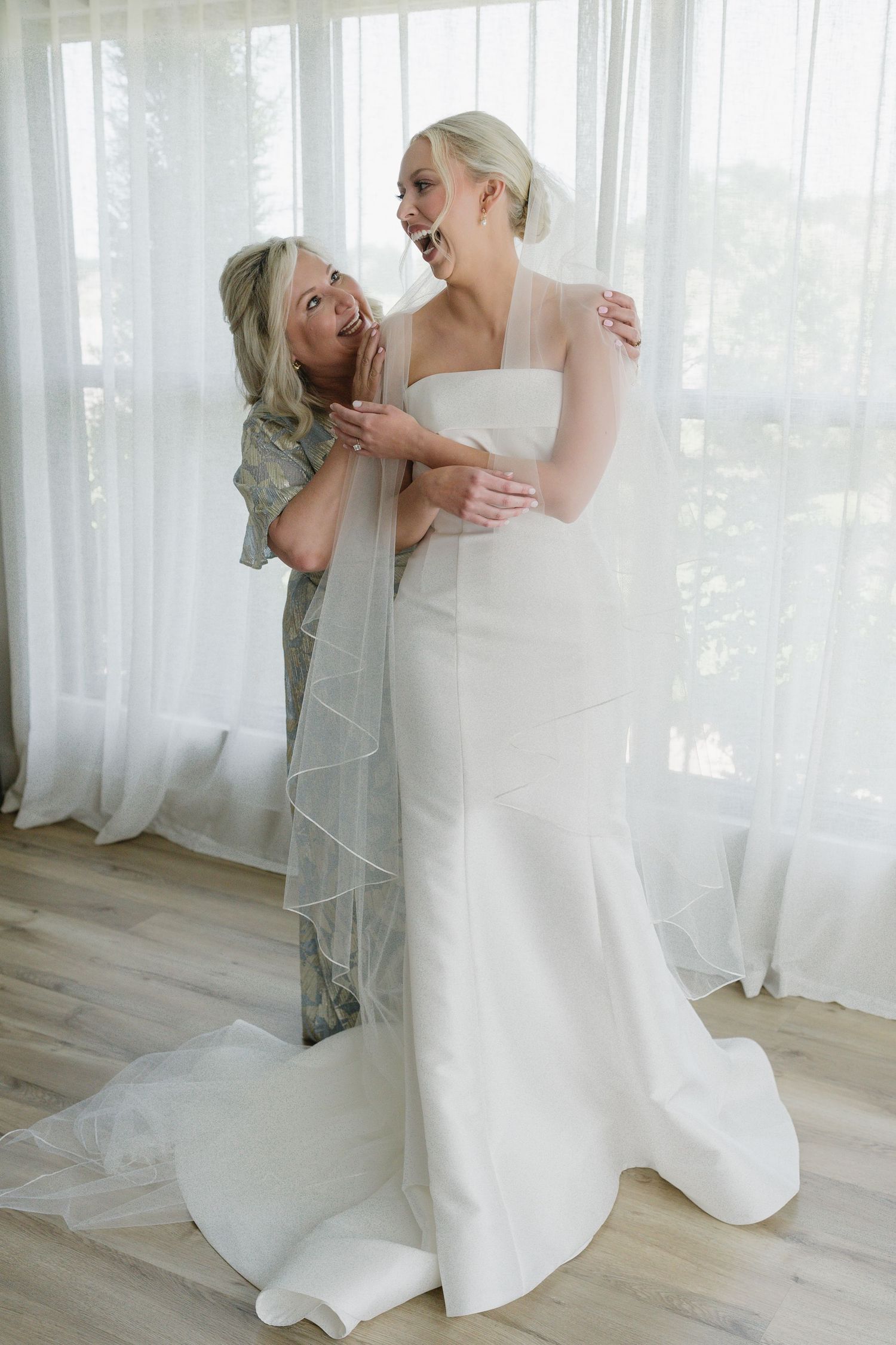 A bride in a white gown and veil shares a joyful moment with a woman helping her dress near sheer curtains.