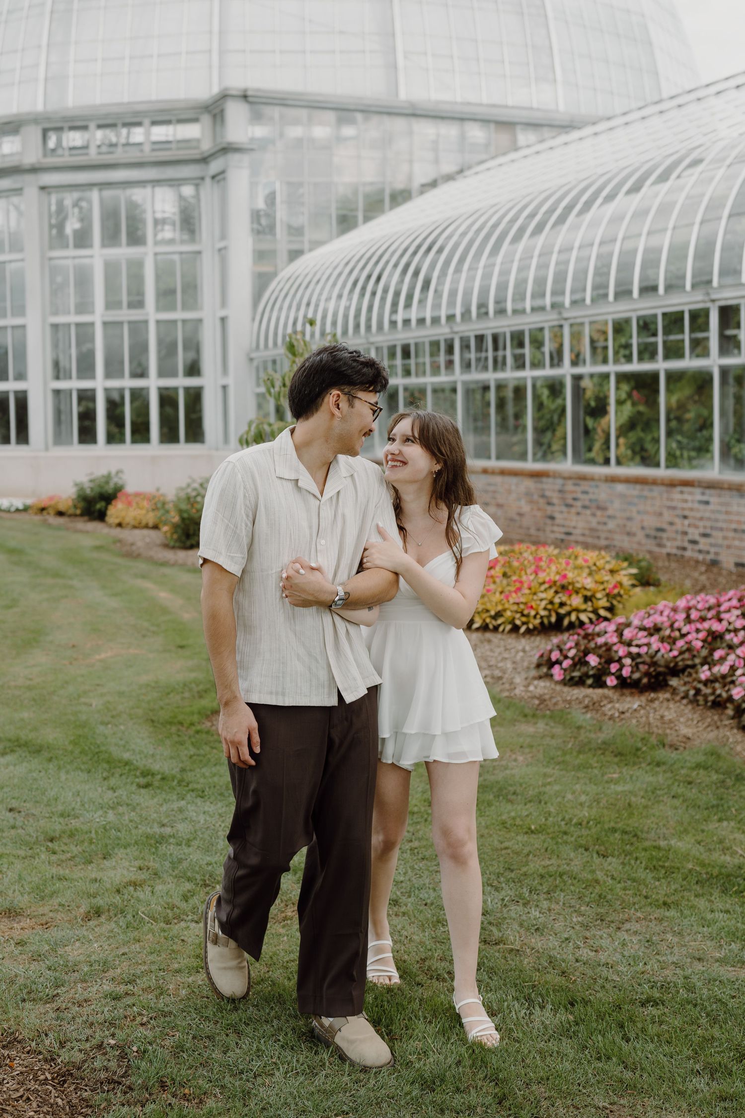 A couple shares a playful interaction on the grass with a glass greenhouse in the background.