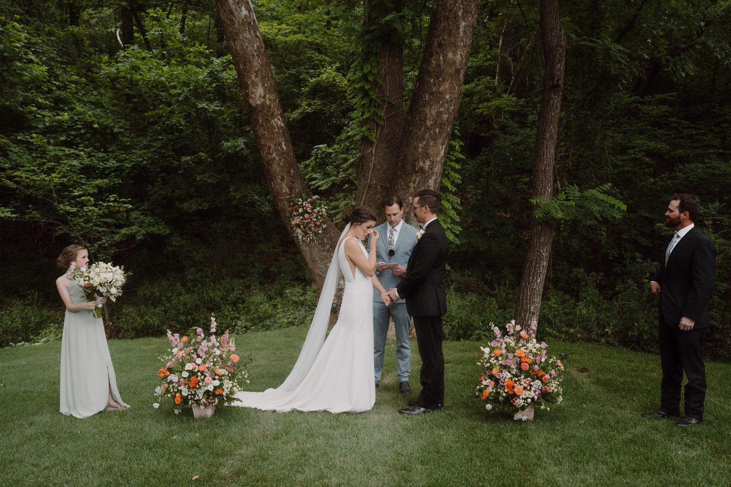 Outdoor wedding ceremony taking place under mature trees with floral arrangements.