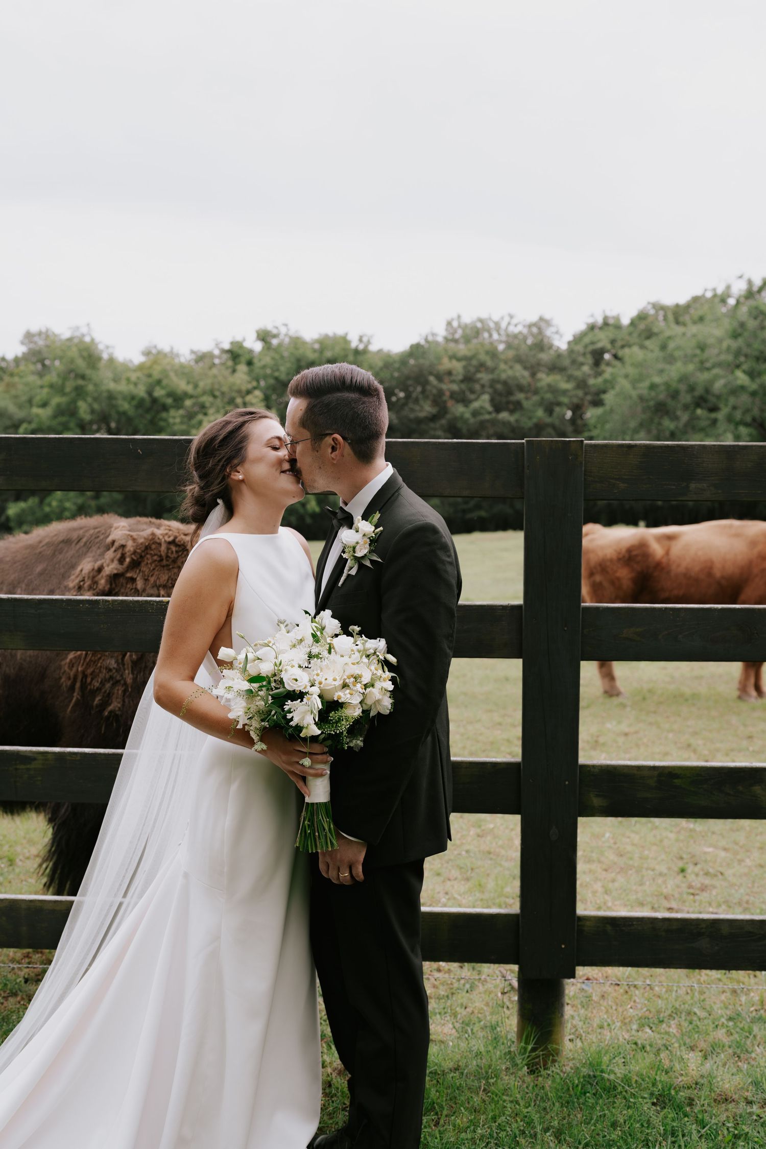 Bride holding white bouquet shares intimate moment with groom by wooden farm fence.