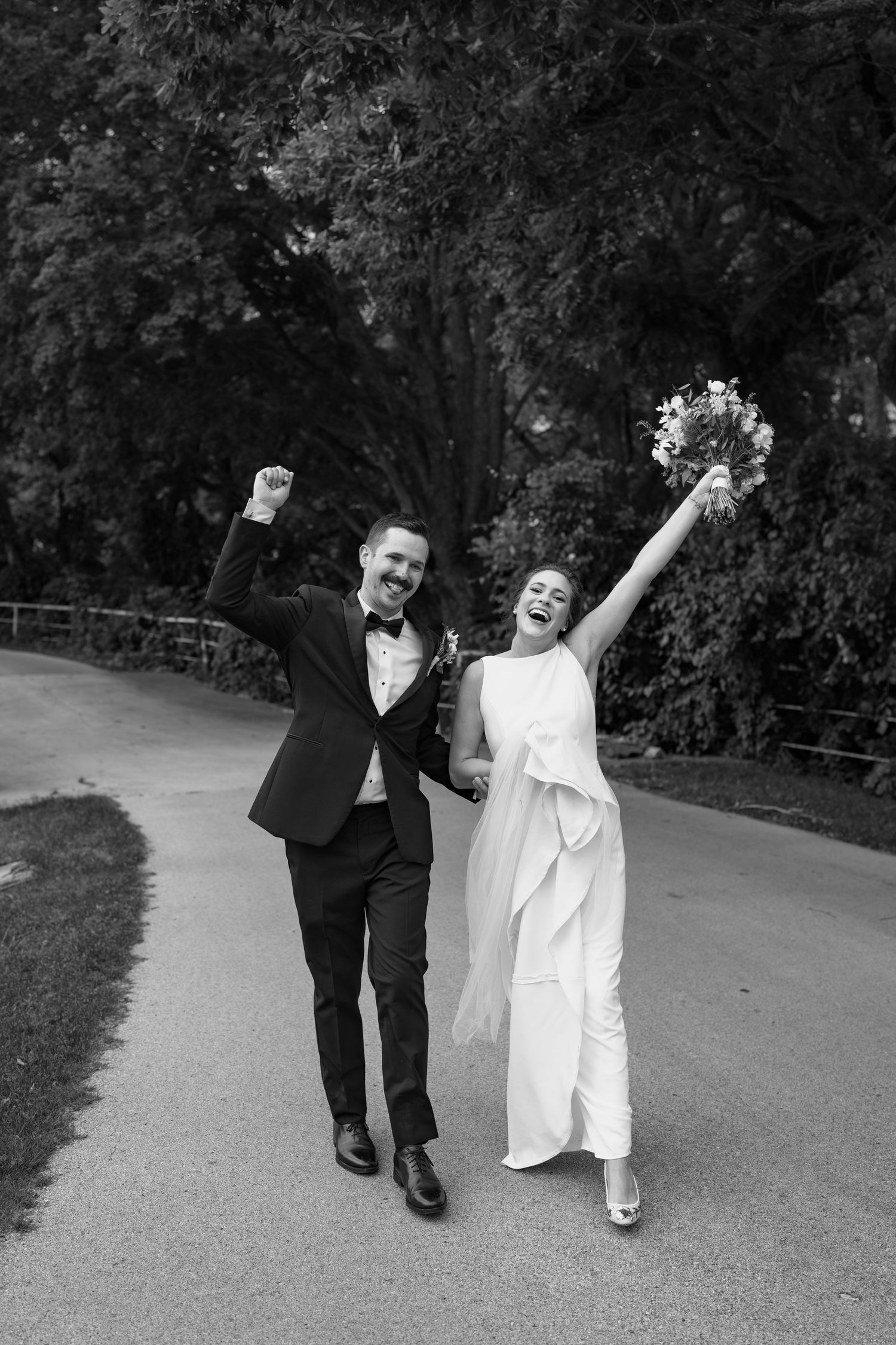 Black and white photo of newlyweds celebrating with energetic poses on a garden path.