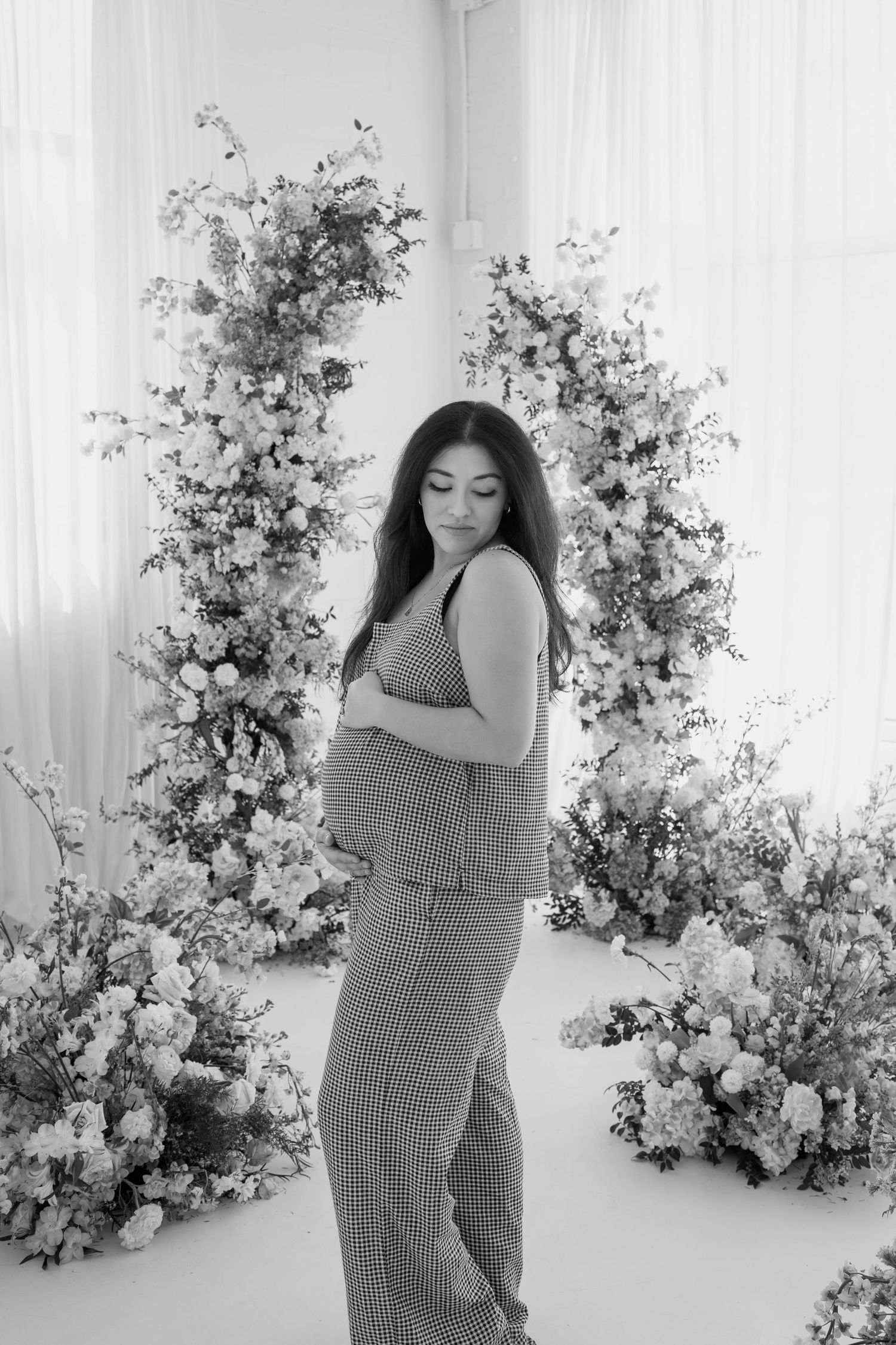Black and white maternity portrait with floral arrangements in minimalist studio setting.