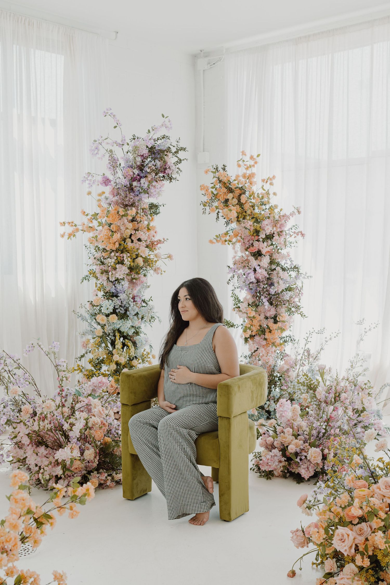 Maternity photo with woman in gingham outfit seated on yellow chair with pink and peach flowers.