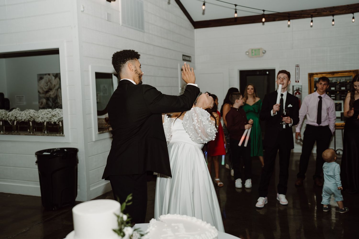 Wedding celebration with guests dancing under string lights in a white venue.