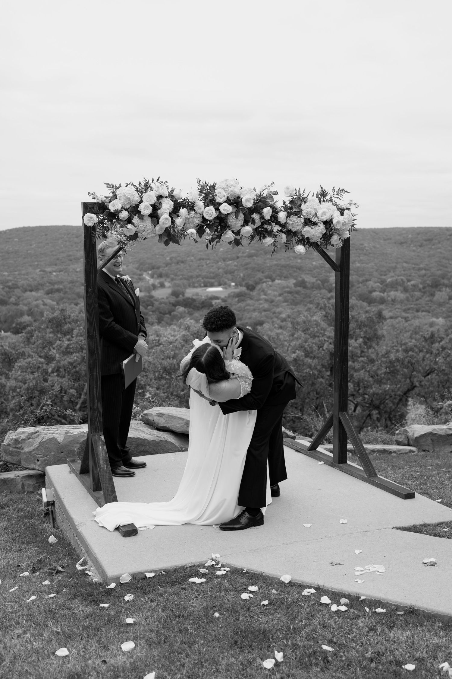 Black and white photo of wedding ceremony on scenic overlook with decorated wooden arch.