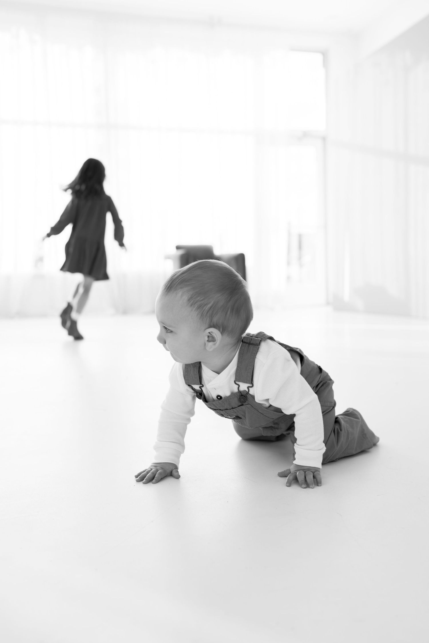 A crawling baby in overalls and a child running in a bright minimalist white room.