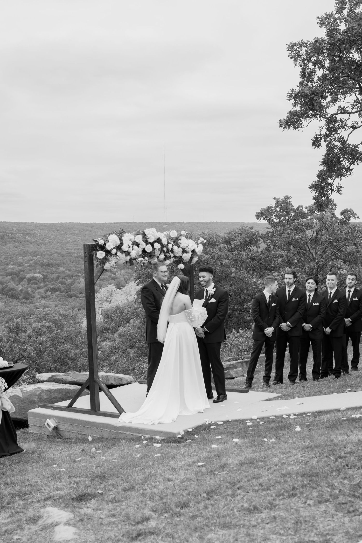 Black and white photo of wedding ceremony on hilltop with floral arch and groomsmen lineup.