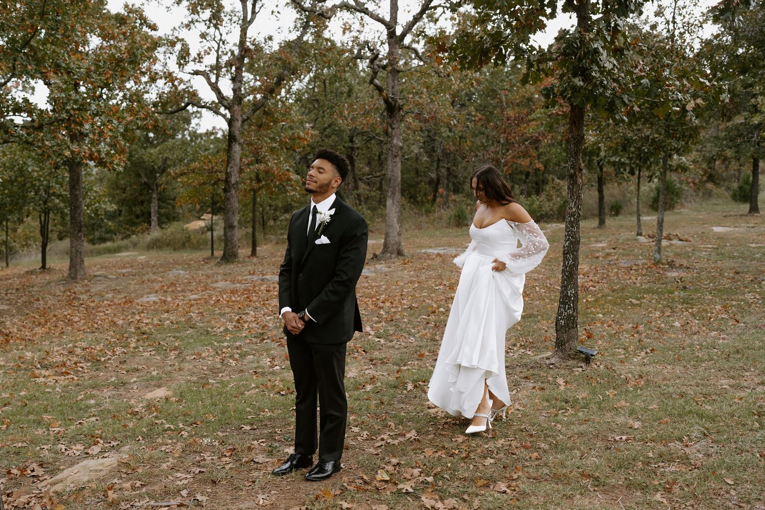 Couple in wedding attire stands apart during first look moment in autumn woodland setting.