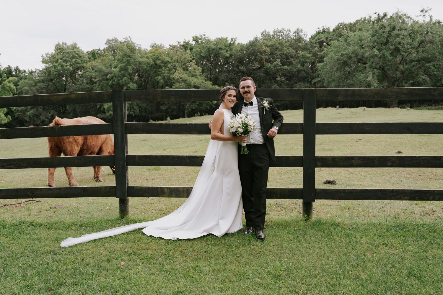 Wedding couple poses in front of a pastoral horse farm with wooden fencing and green fields.