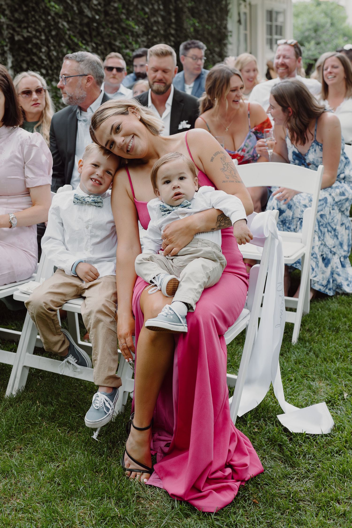 Wedding guests seated in white chairs on grass with children in formal attire.