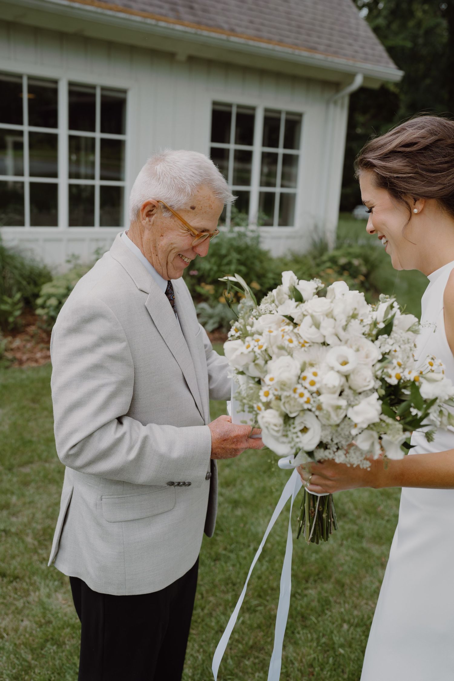 Wedding first look scene with white bouquet and light gray suit against white building.