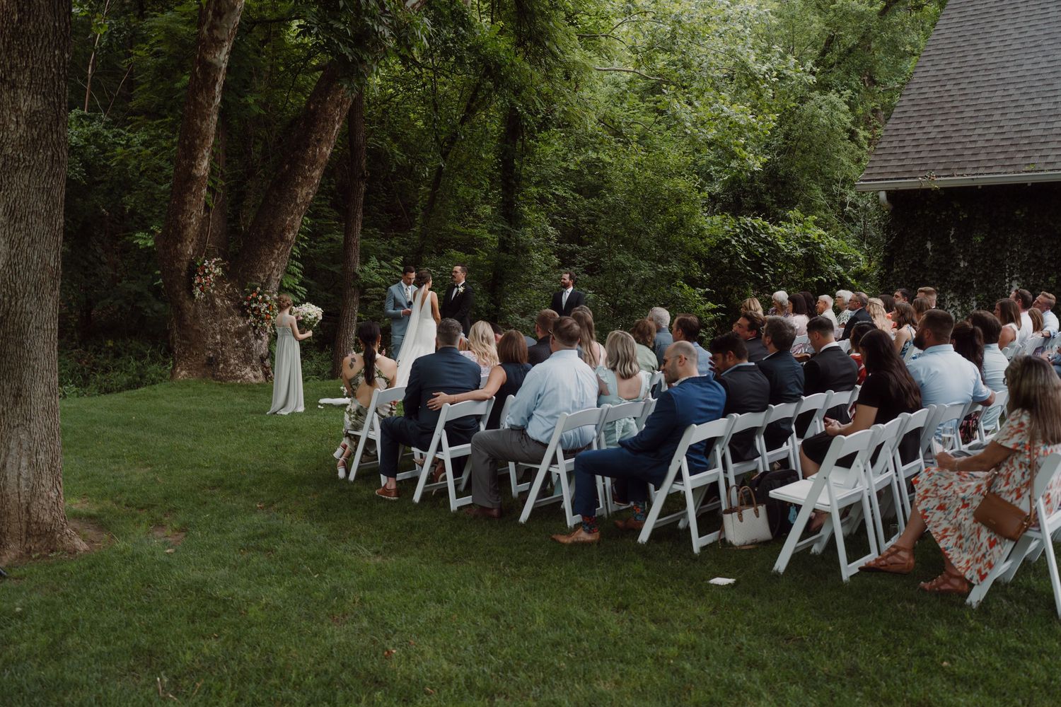 Wedding guests seated in rows of white chairs during an outdoor forest ceremony.