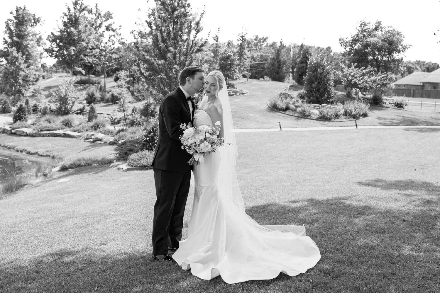 Black and white photo of a bride and groom sharing a kiss on a lush green lawn with trees in the background.