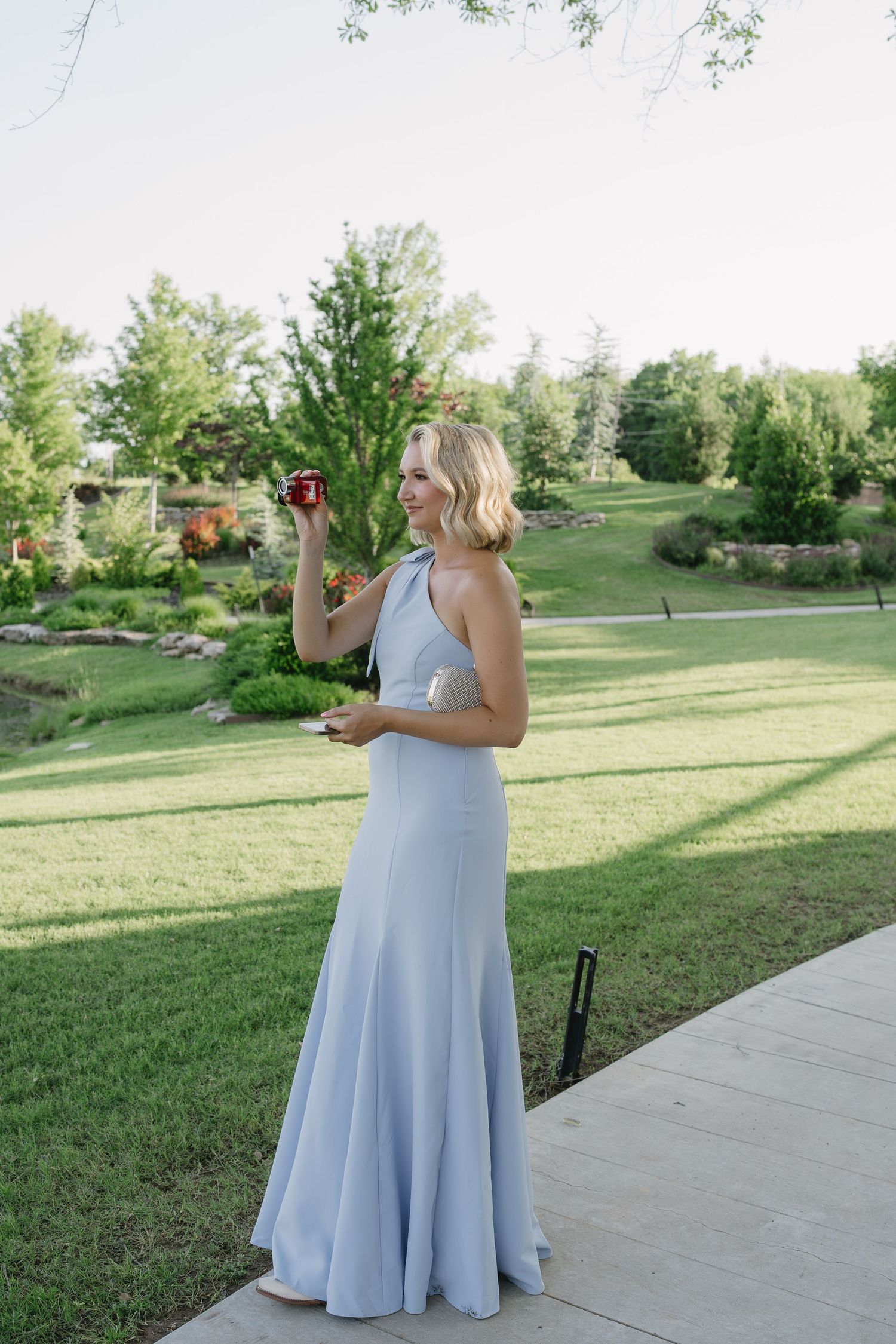 Blonde woman in light blue halter gown holding red drink outdoors at garden wedding reception.