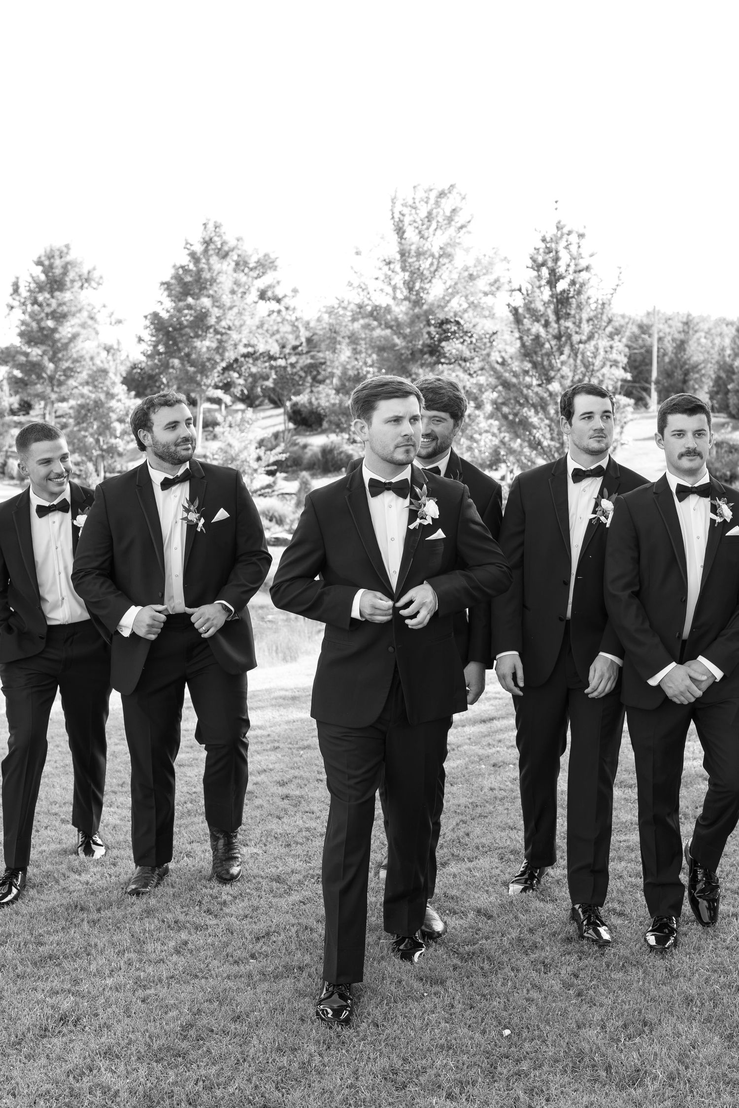 Black and white photo of a groom and groomsmen in tuxedos with bow ties walking outdoors at a wedding.