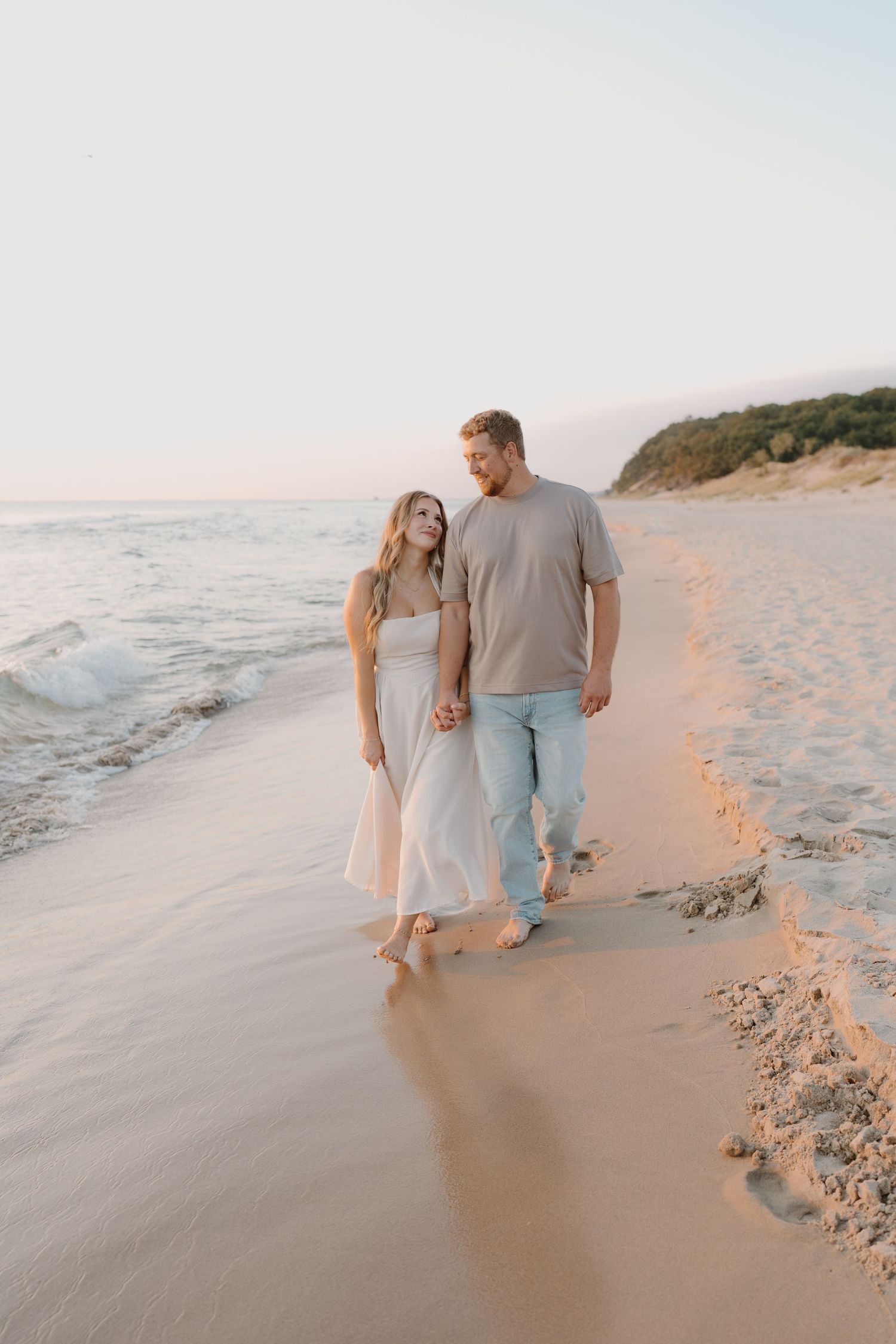A couple walks hand in hand along a serene beach at sunset with gentle waves rolling onto the shore.