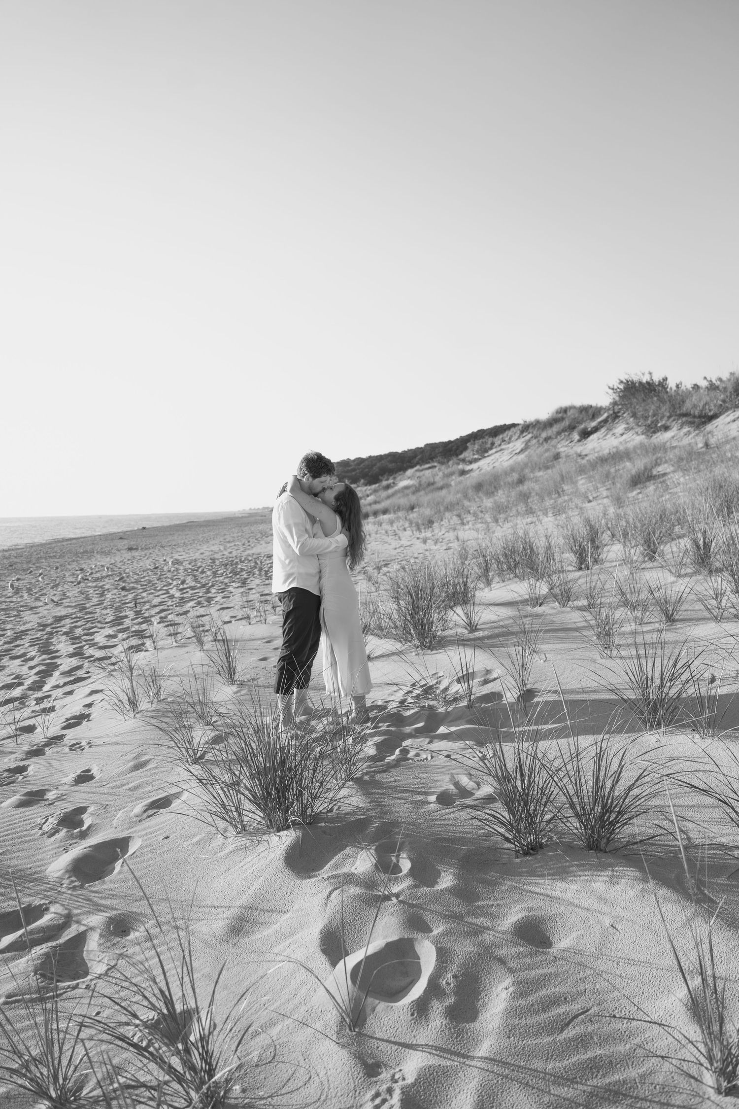A black and white beach portrait showing a couple's embrace among windswept dunes and beach grass.