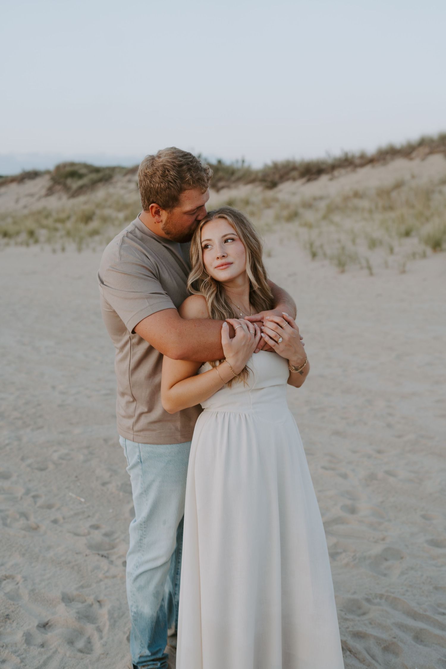 A romantic embrace captured during a coastal photoshoot with sand dunes in the background.