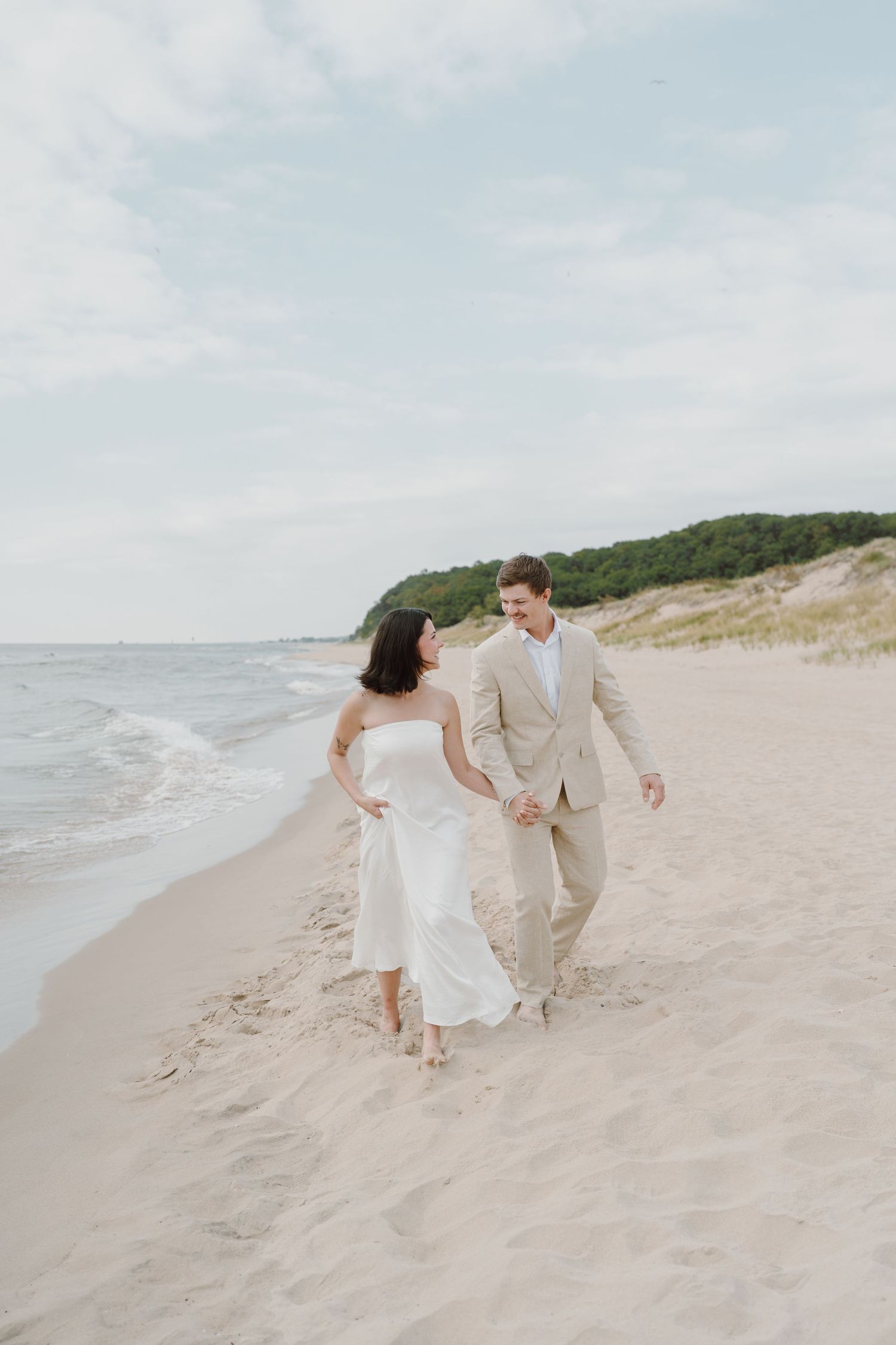 A couple in light-colored clothing walks together on a scenic beach with dunes.