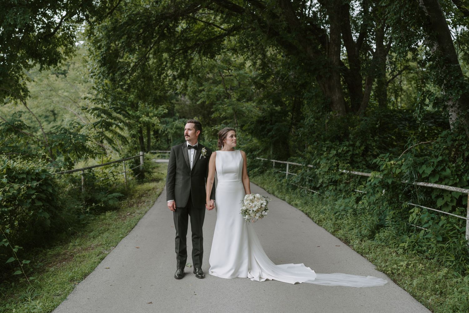 A couple in wedding attire strolls down a scenic path surrounded by green trees and natural scenery.