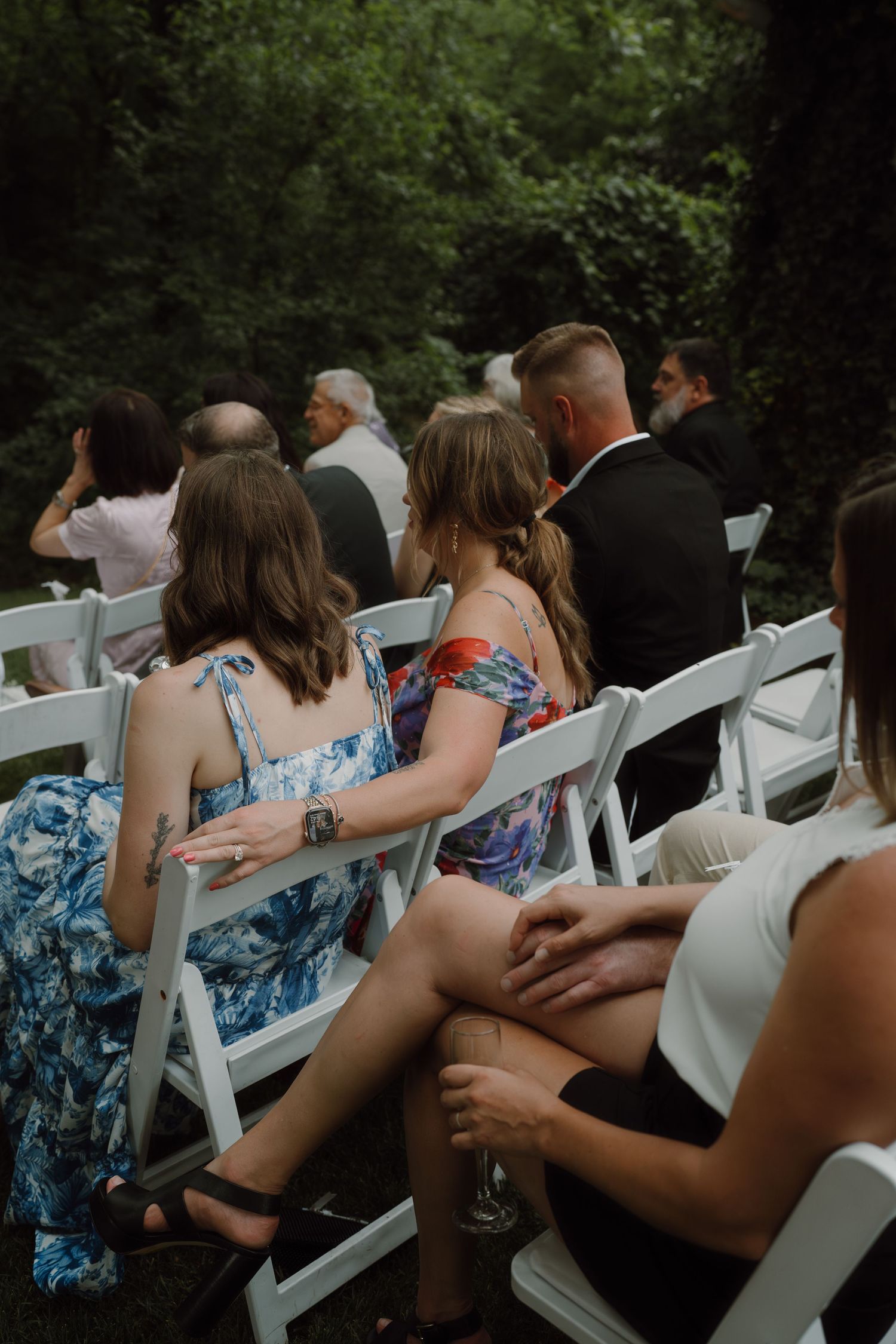 Wedding guests seated in white chairs during an outdoor ceremony in a wooded setting.