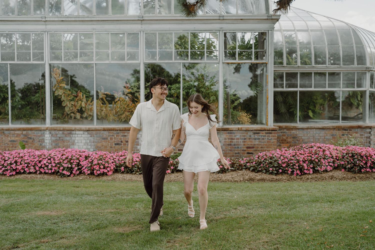 Two people walk hand in hand on grass near a glass greenhouse structure.