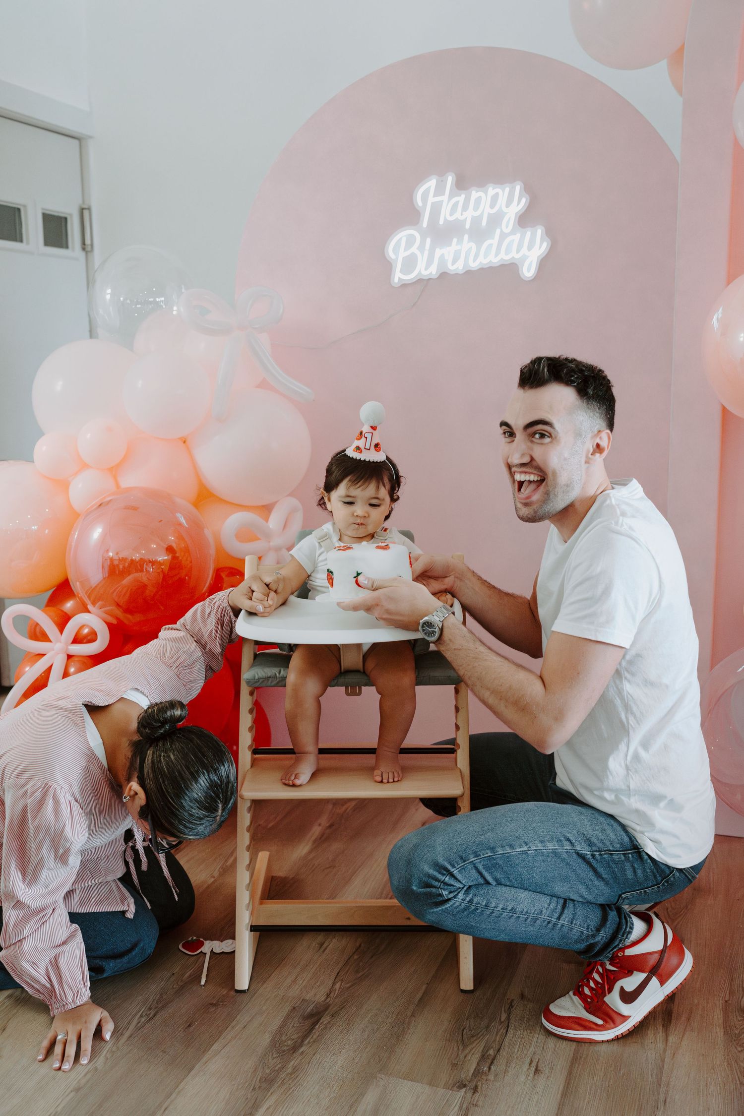 Birthday party setup with pink balloon wall and neon sign as family shares special moment.