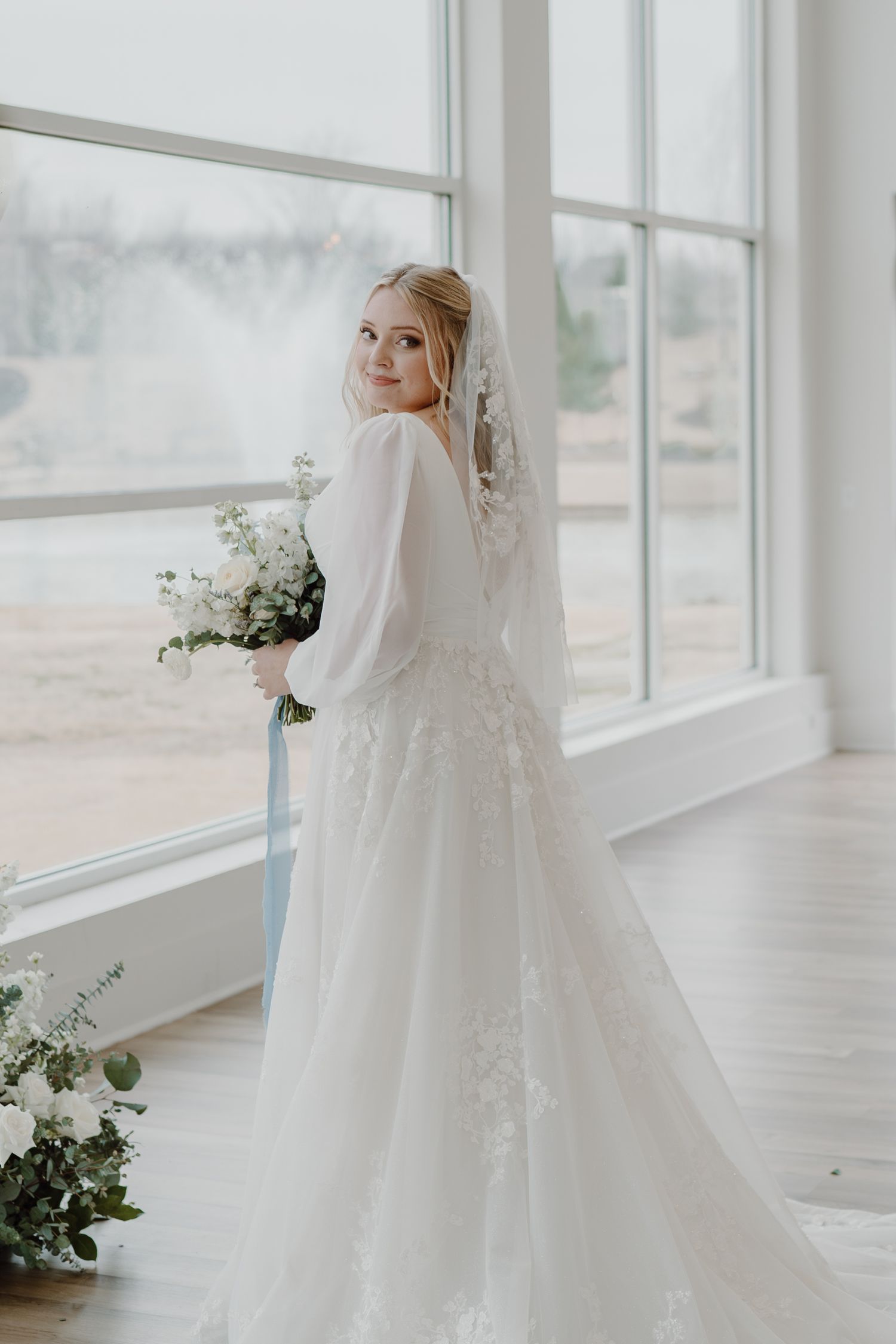 Elegant bride in long sleeve white wedding gown poses by large windows with floral arrangements.