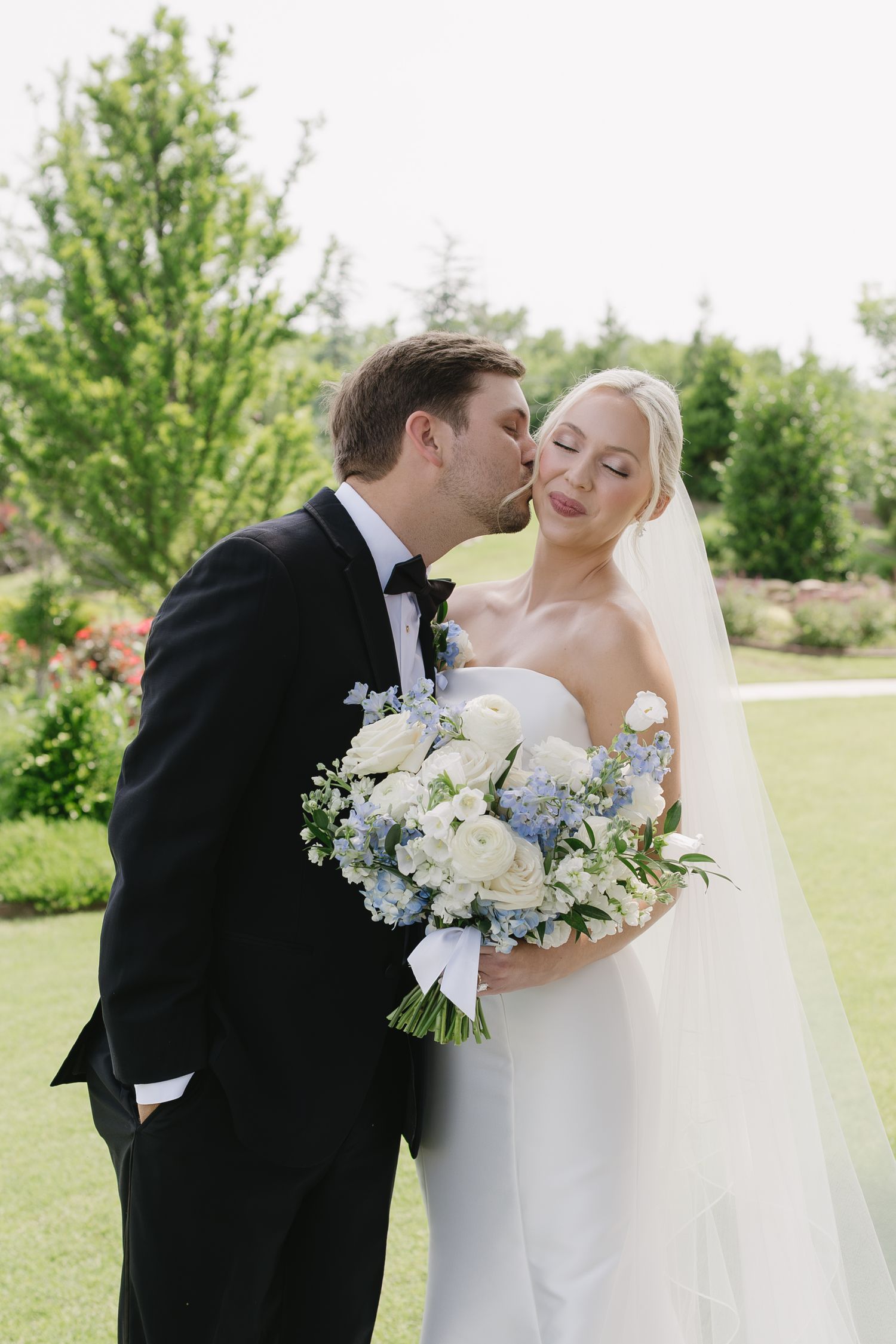 Groom in black tuxedo kisses bride holding white and blue bouquet in lush garden wedding photo.