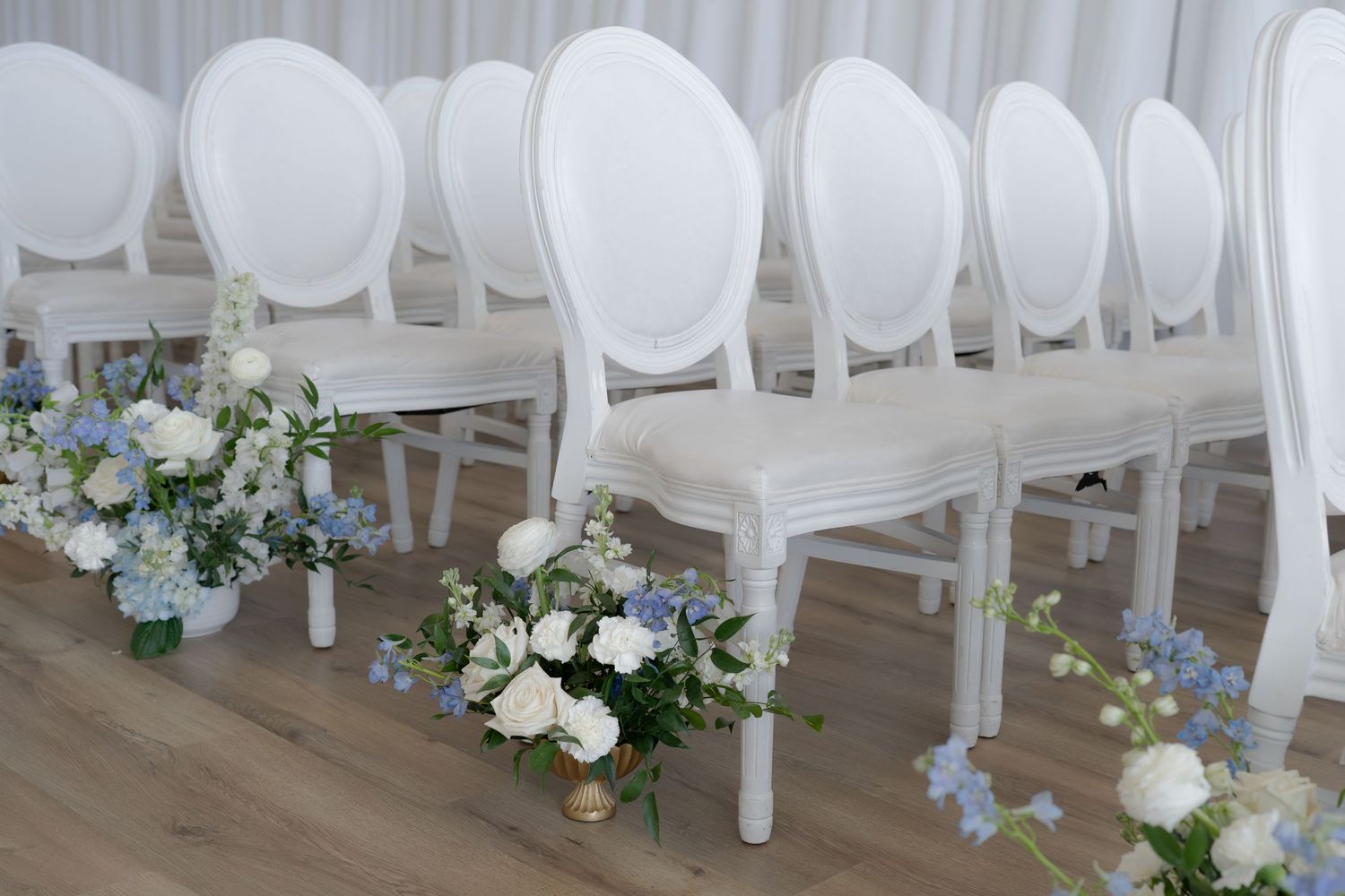 Elegant white Louis XVI chairs arranged in rows with blue and white floral arrangements on a wooden floor.