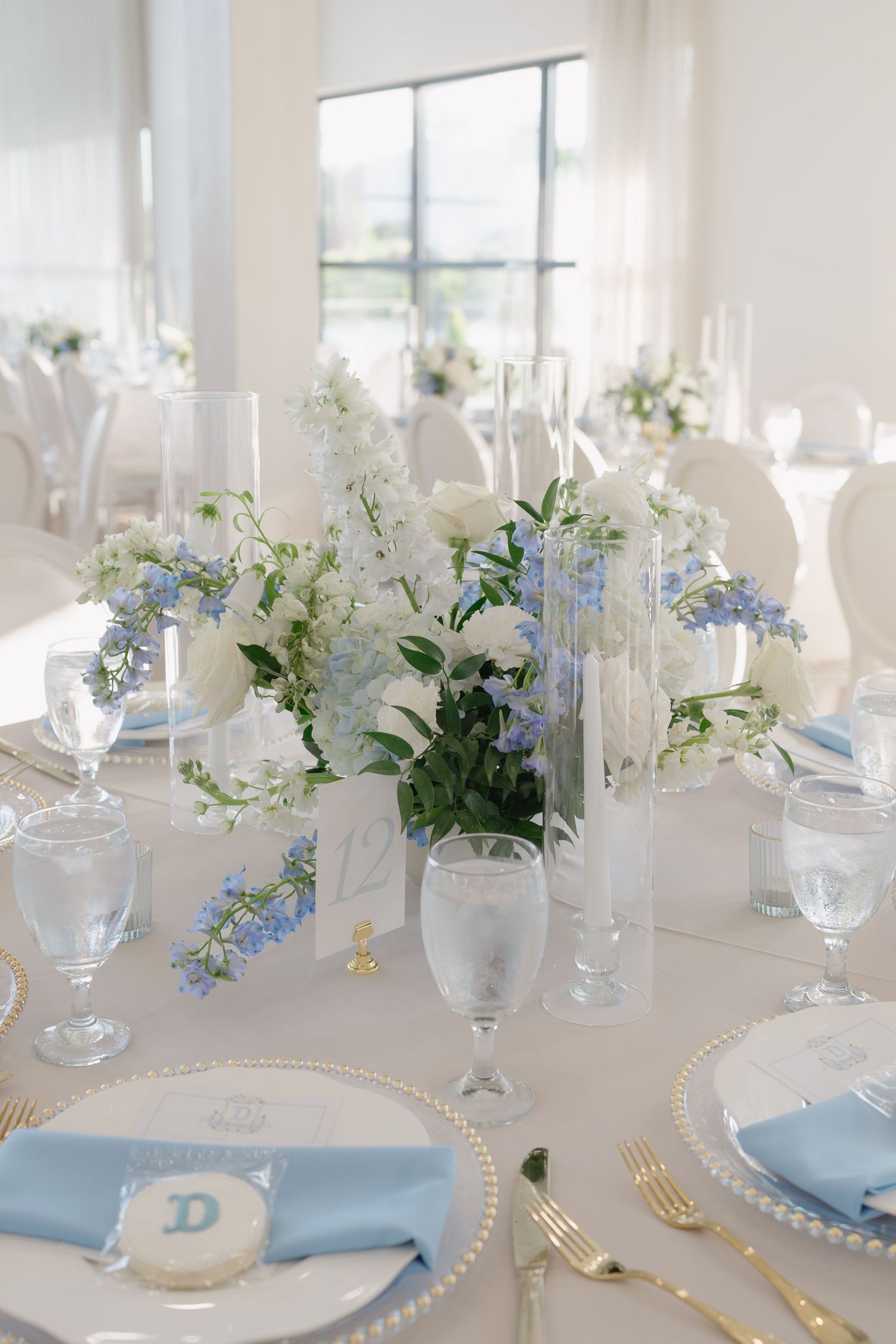 Elegant wedding reception table with blue and white floral centerpiece, gold-rimmed chargers, and light blue linen napkins.