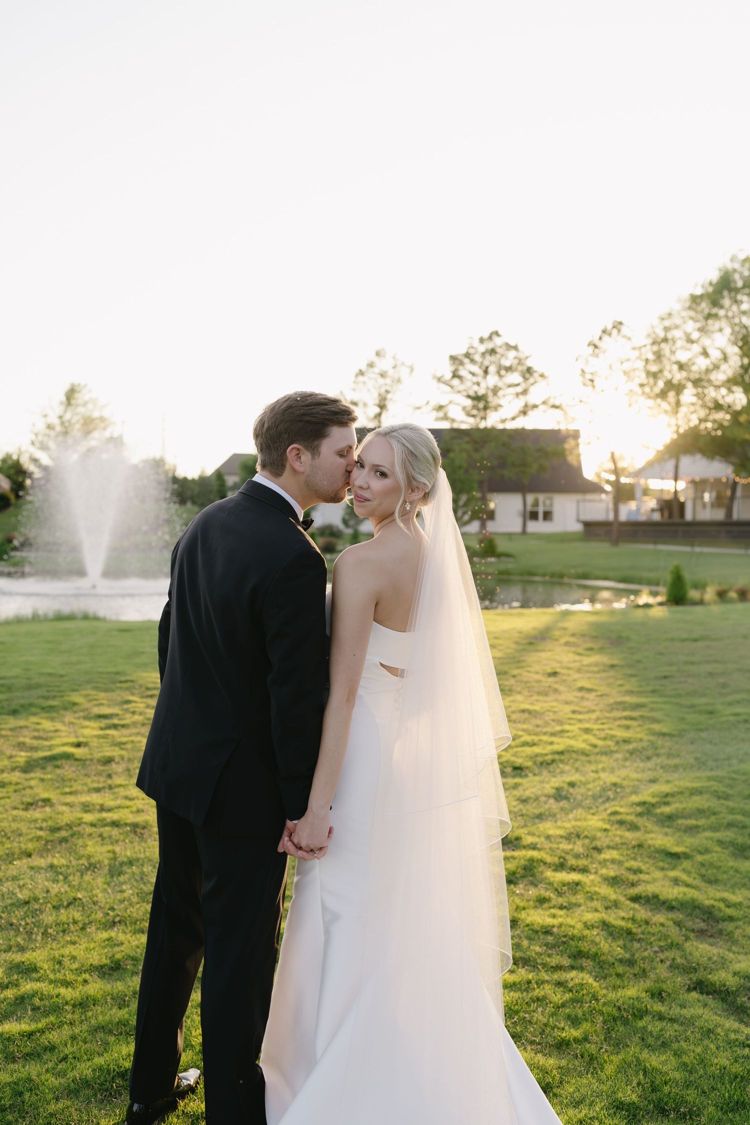 A groom in a black suit kisses his bride in a white gown and veil outdoors on a green lawn at sunset.