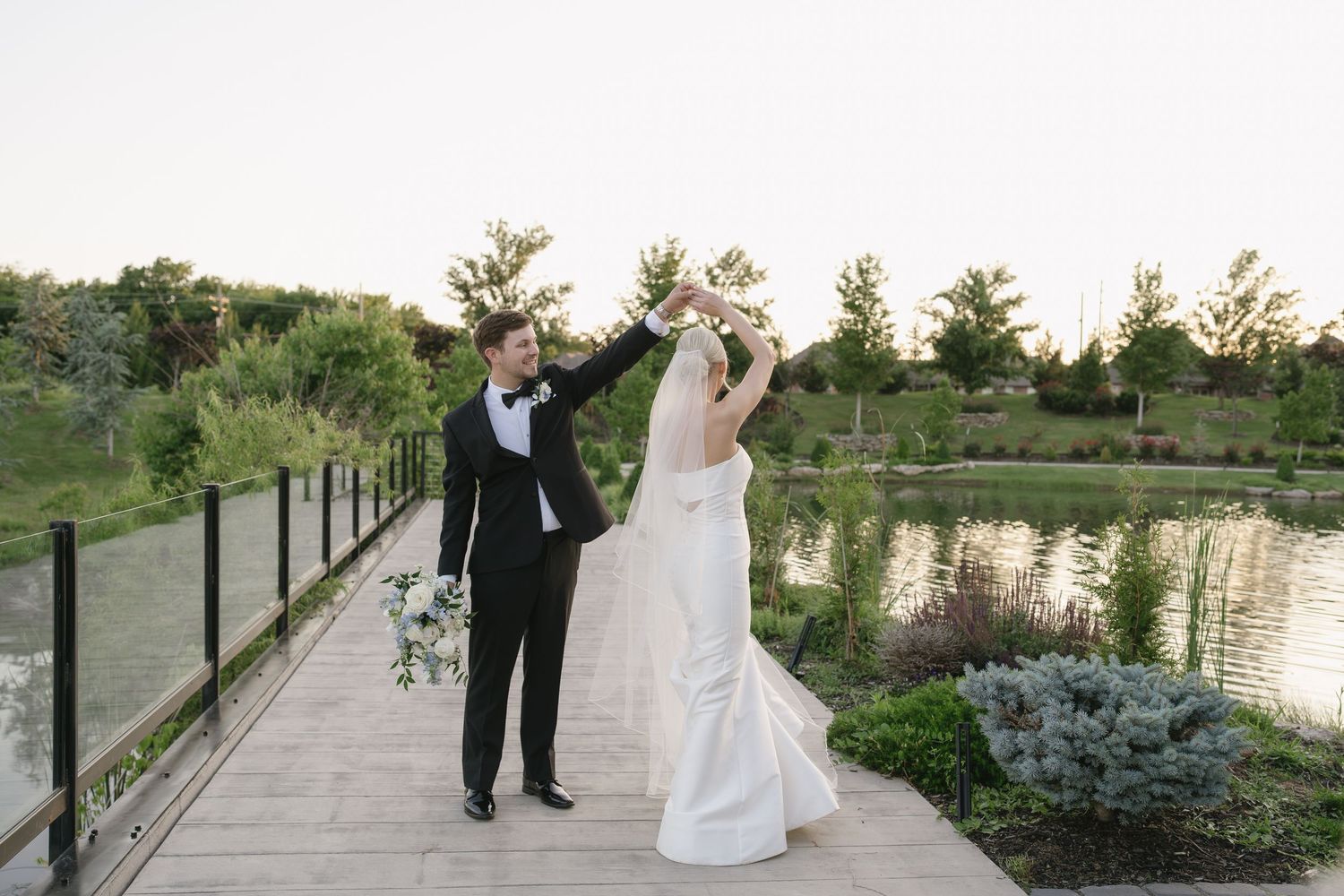 Bride and groom dancing on a wooden bridge by a pond, surrounded by lush greenery, in elegant wedding attire.