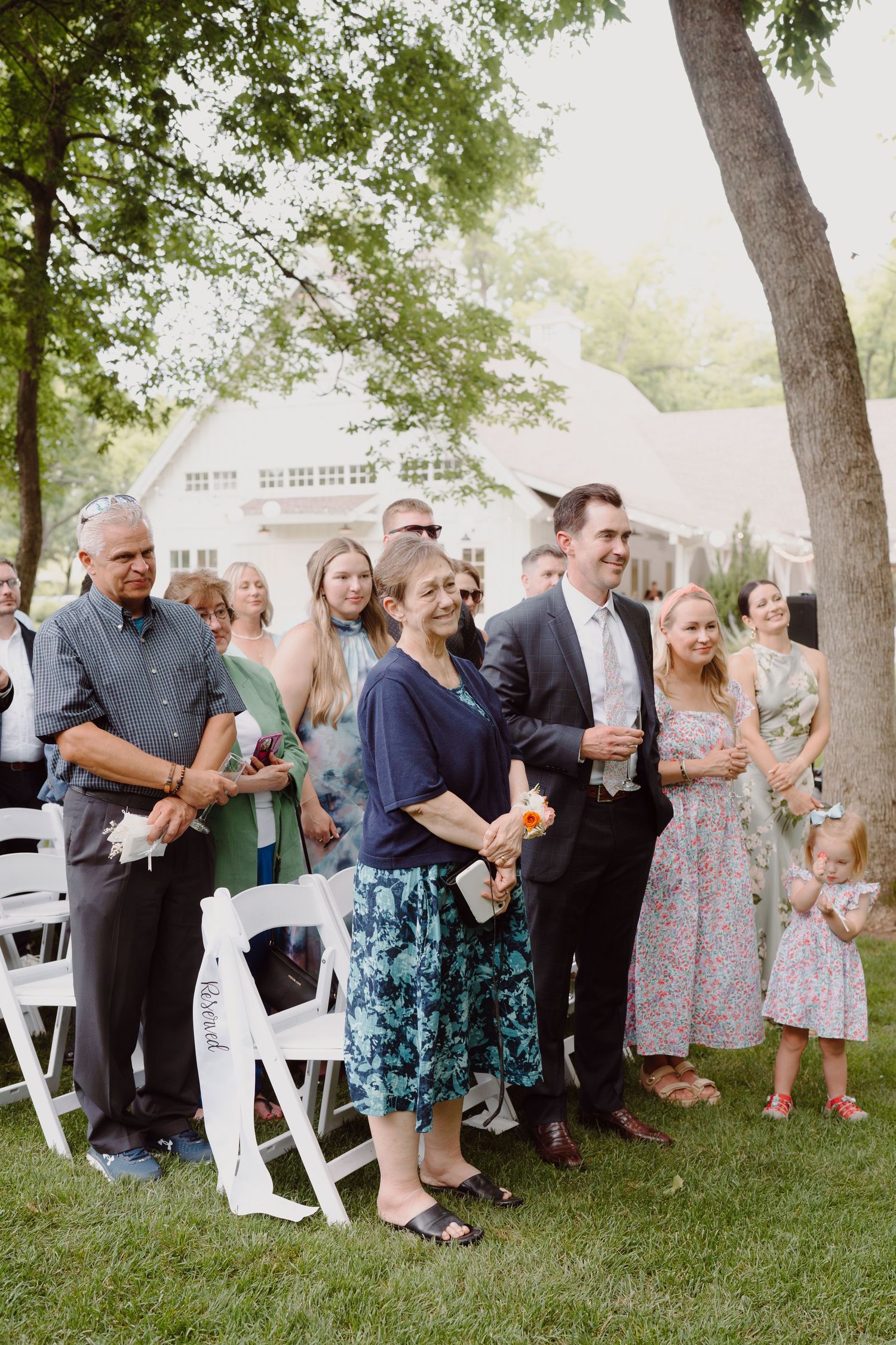 Wedding guests gather on a lawn near white folding chairs during an outdoor celebration.
