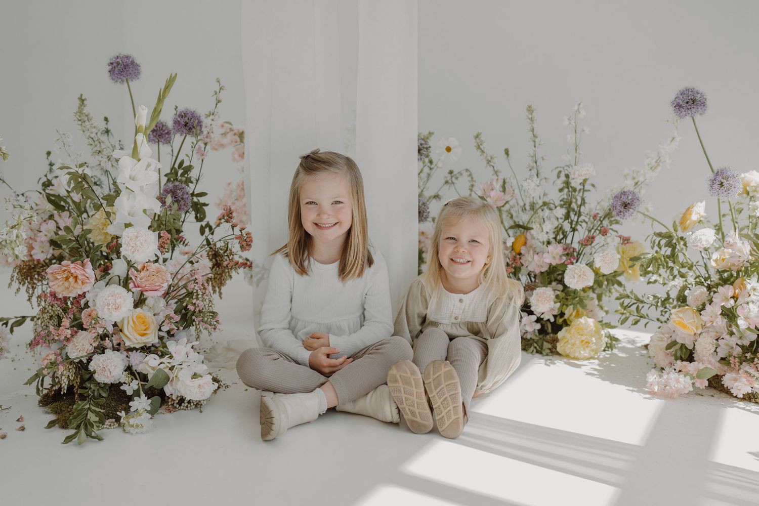 Bright studio portrait with siblings sitting together surrounded by pastel flower arrangements.