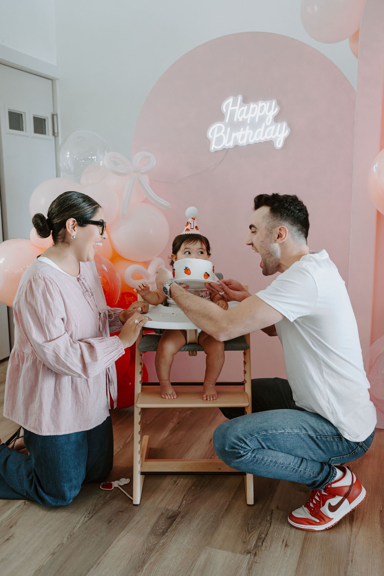 Intimate family moment during birthday celebration with pink balloon wall and decorative lighting.
