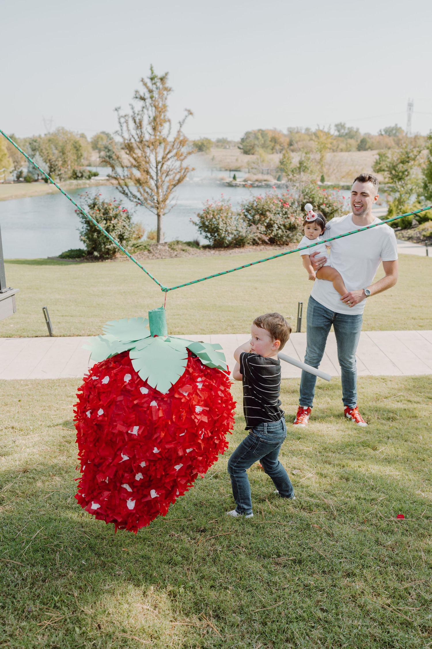 A bright red heart piñata is being swung on a rope in a park setting.
