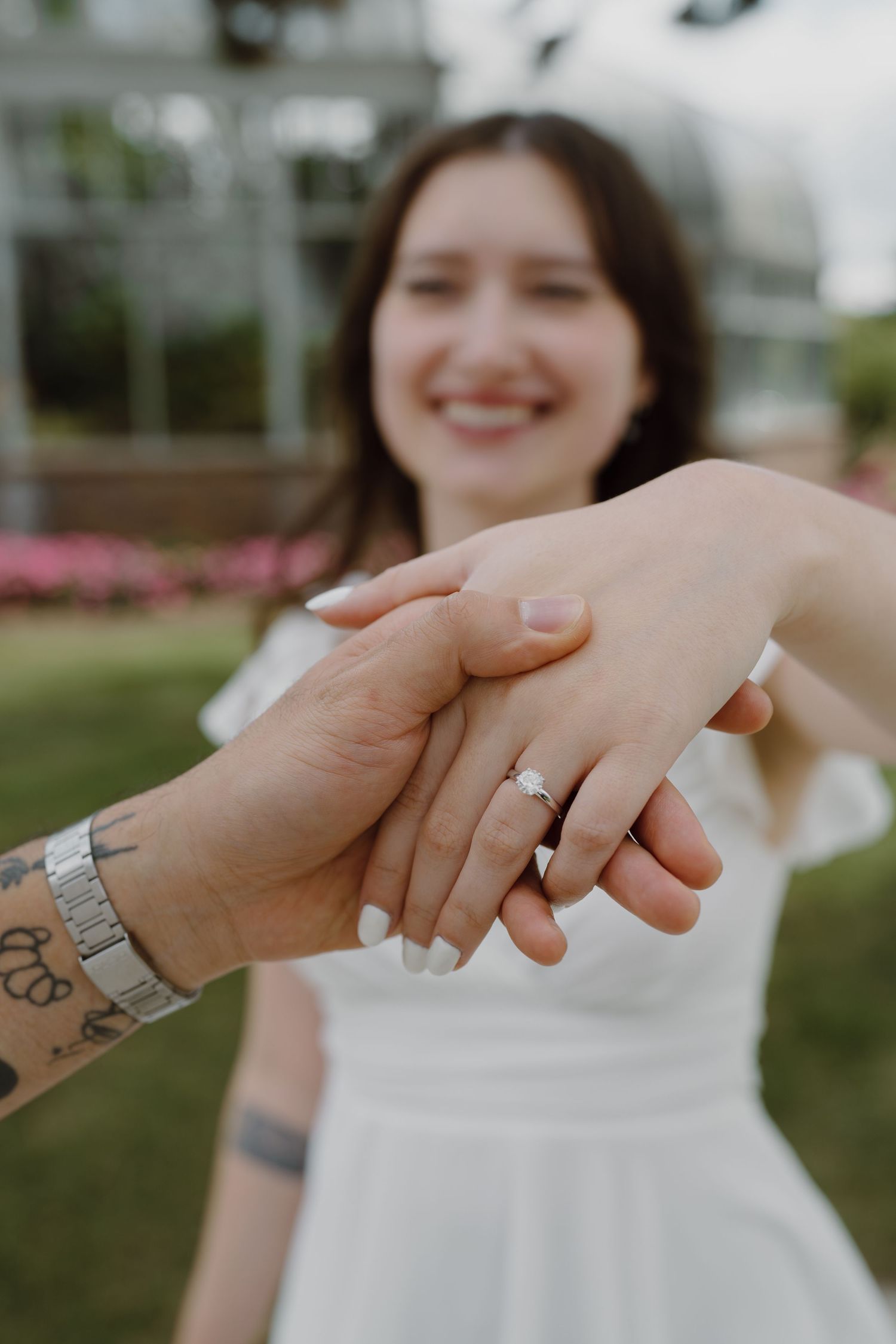 Close-up of hands with rings and tattoos touching against a blurred greenhouse background.