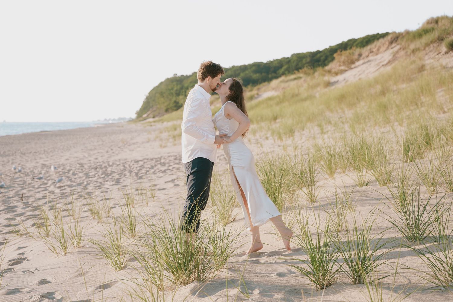 A loving couple in light colored clothing embraces on a sandy beach near grass-covered dunes.