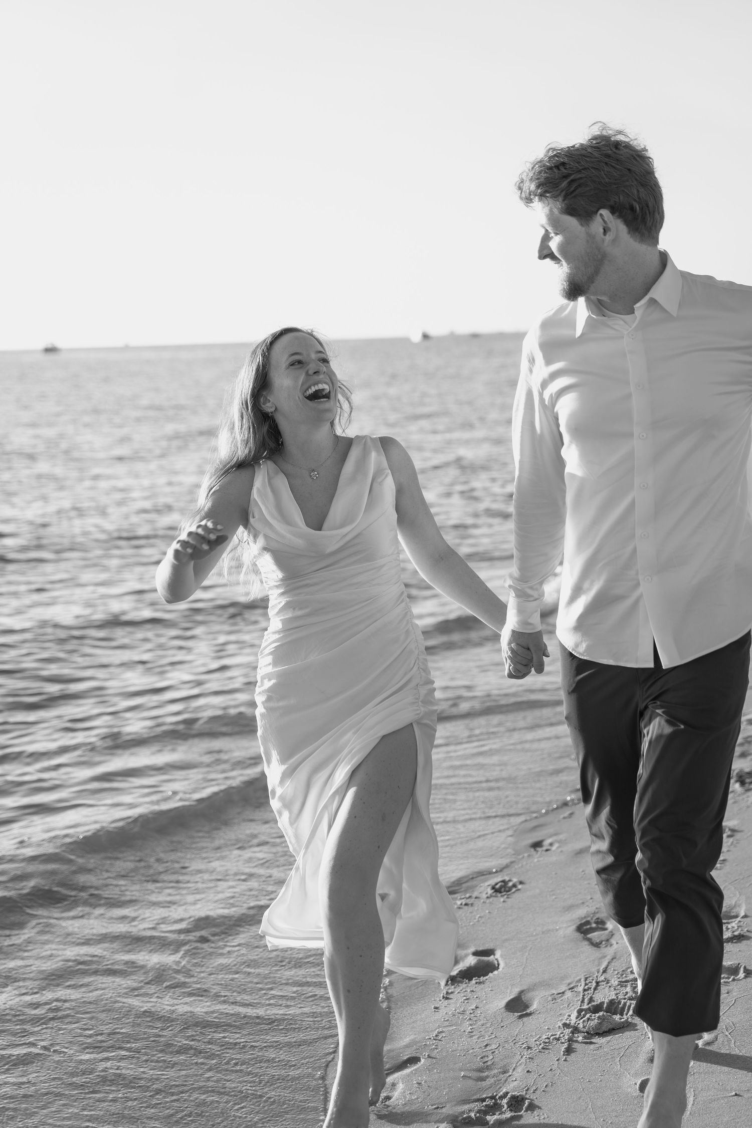 A joyful couple laughs while running together on a beach at sunset.