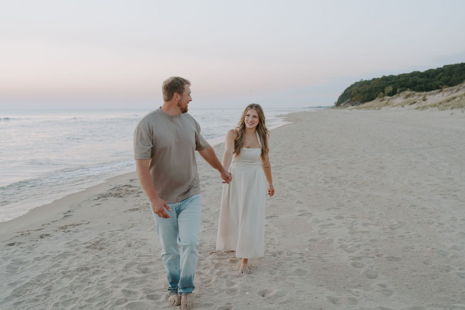 A couple walks hand in hand along a serene beach at sunset with ocean waves in the background.